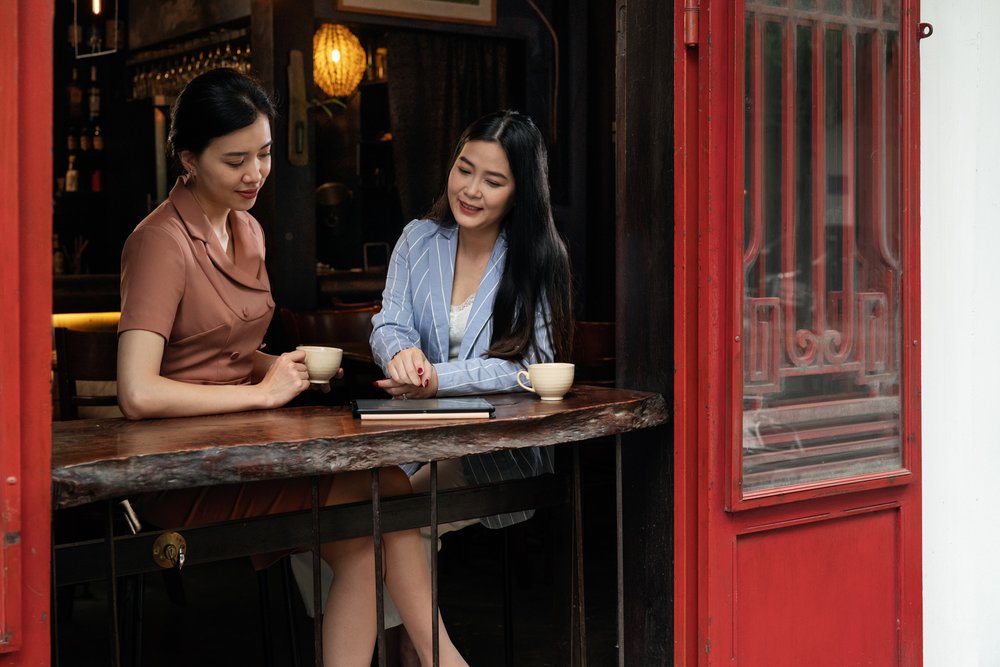  Female financial professionals in discussion with tablet device while sitting at open window in artistic cafe. 