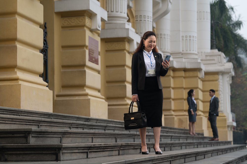  Female regulatory officer checking her phone on steps of financial building. 