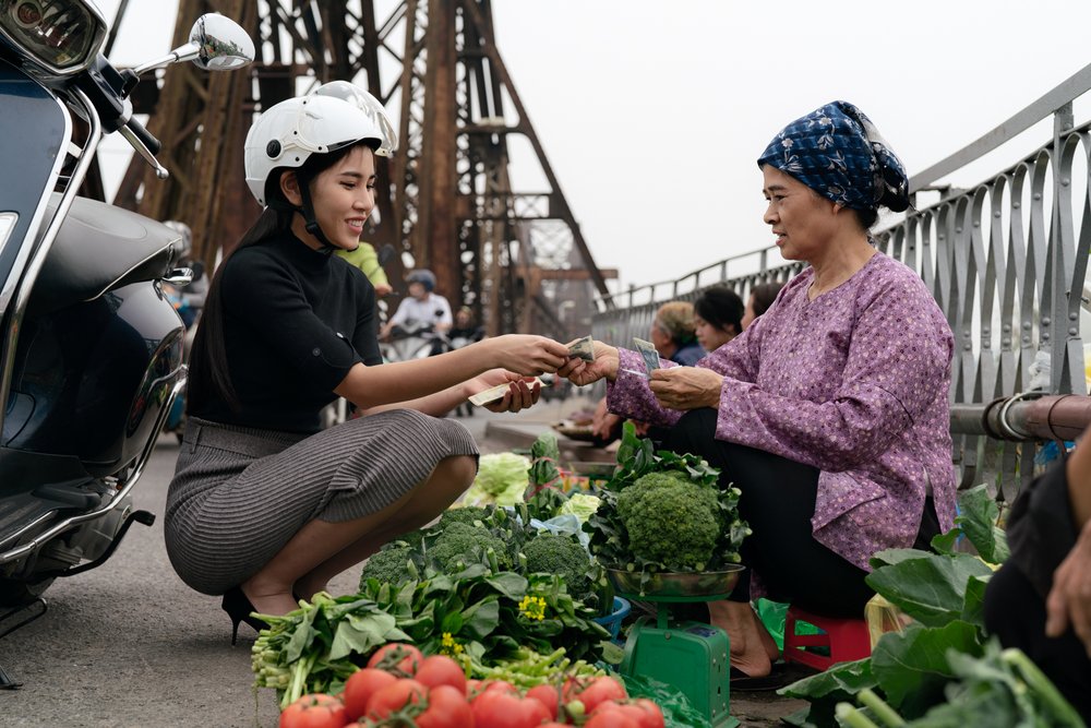 City girl making a cash payment as she purchases vegtables from a farmer on Long Bien Bridge, Hanoi as traffic passes. 