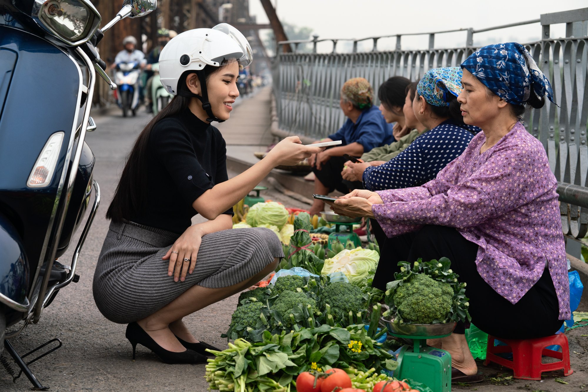  City girl making a phone to phone payment as she purchases vegtables from a farmer on Long Bien Bridge, Hanoi as traffic passes. 