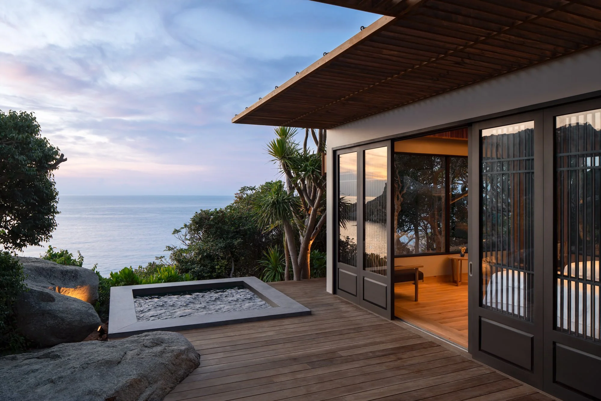  Amanoi Ocean Pool Residence outdoor jacuzzi on a wooden terrace overlooking the ocean, surrounded by tropical vegetation and granite boulders, with the private spa interior visible at dawn. 