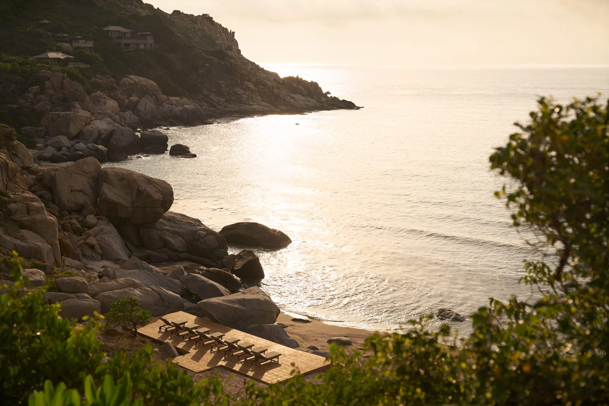  Amanoi Ocean Pool Residence overlooking its private beach and rocky coastline at sunrise, with sunlit sea, loungers and surrounding tropical vegetation. 