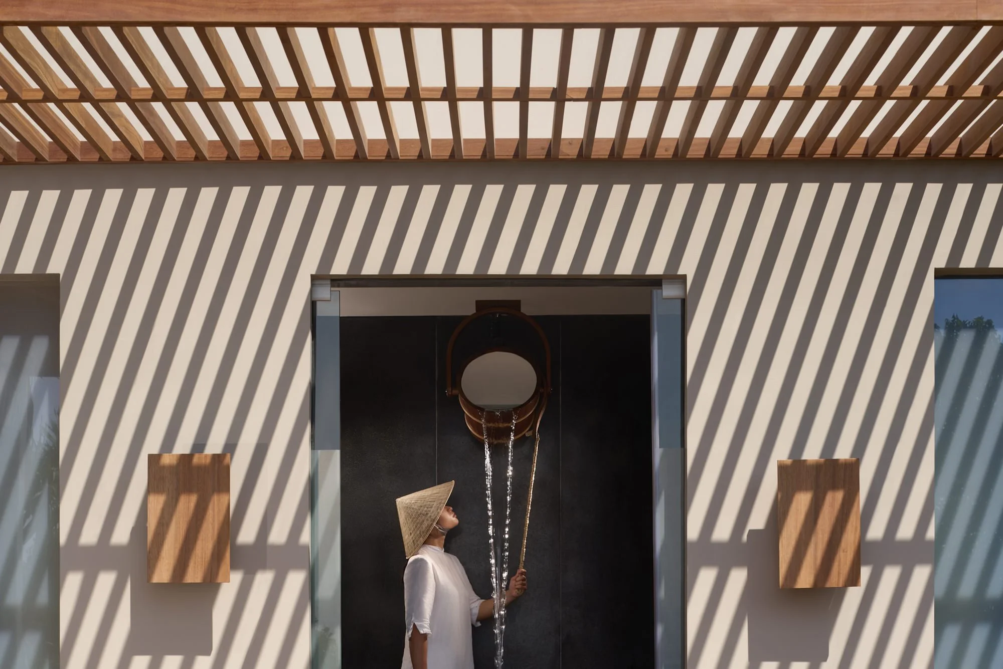  An Amanoi staff member in a traditional conical hat pulls the cord of an ice-bucket shower in the private spa pavilion at Amanoi Ocean Pool Residence, surrounded by lush vegetation and granite boulders. 