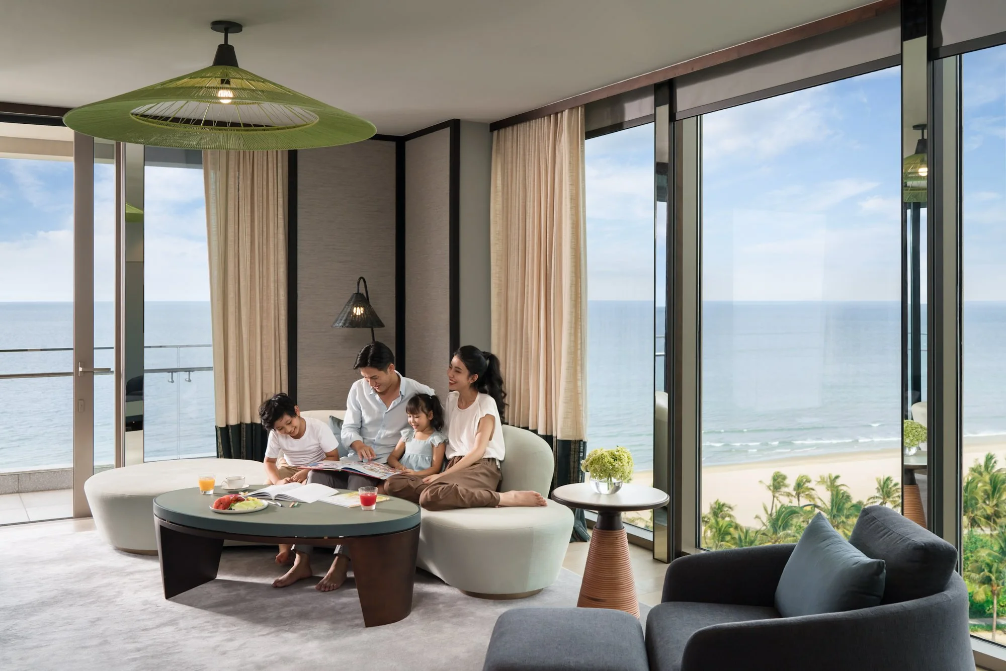  Family relaxing together in a spacious ocean-view room at New World Hoiana Beach Resort, with floor-to-ceiling windows overlooking the beach and sea. 
