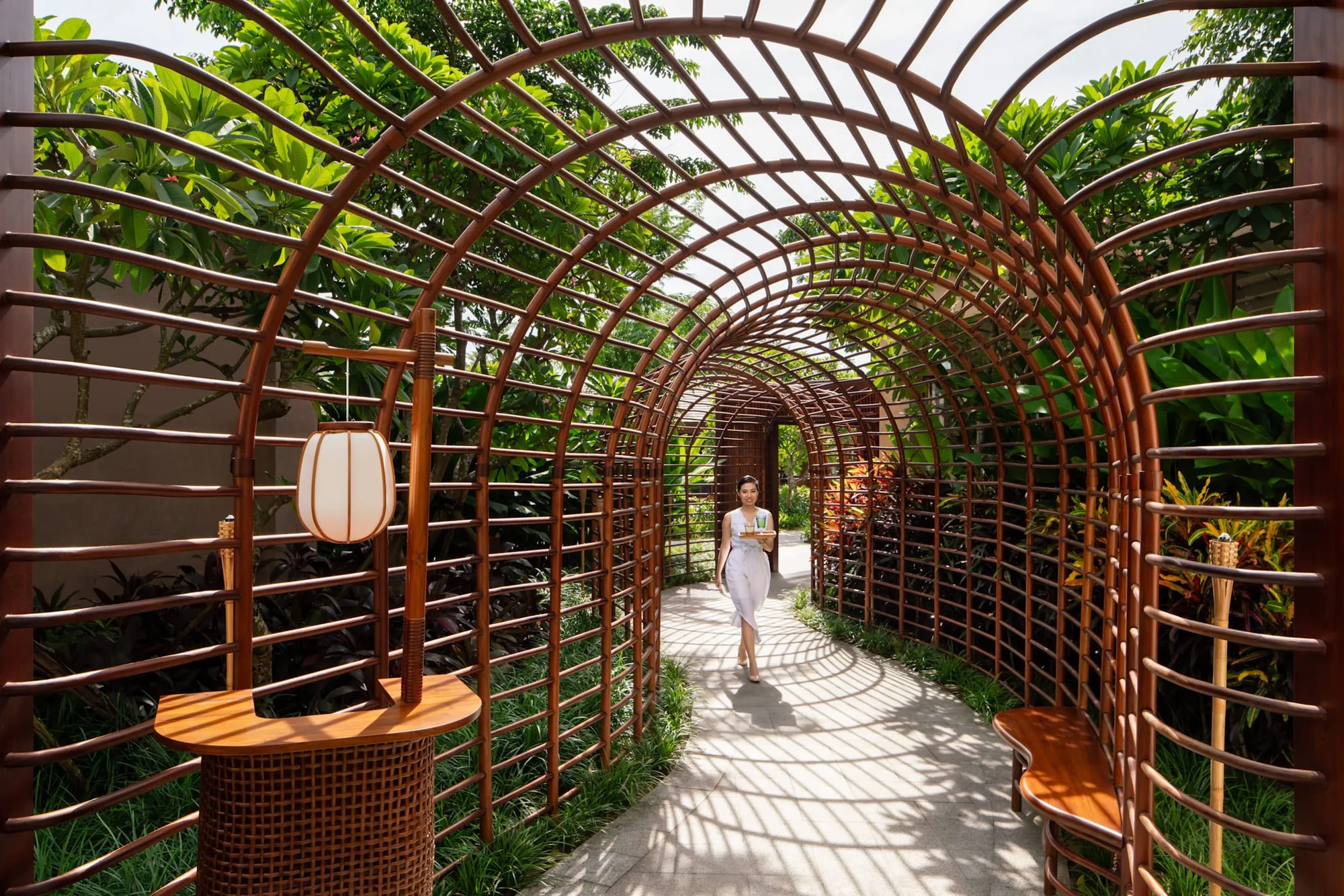  NOX Beach Club waitress walking through a bamboo-covered garden walkway at New World Hoiana Beach Resort, surrounded by lush tropical landscaping. 