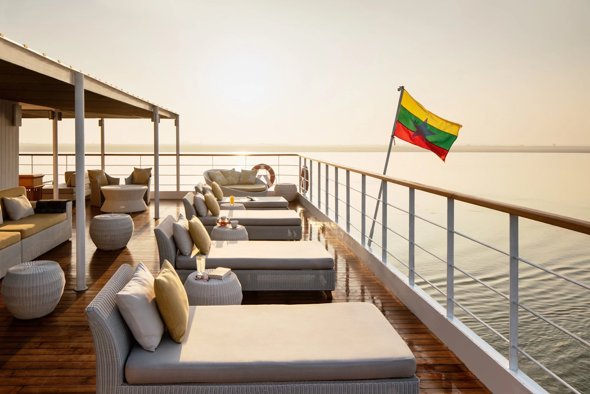  The Strand Cruise sundeck in Myanmar at sunset, with sun loungers, sofas and armchairs overlooking the Irrawaddy River and surrounding landscape. 