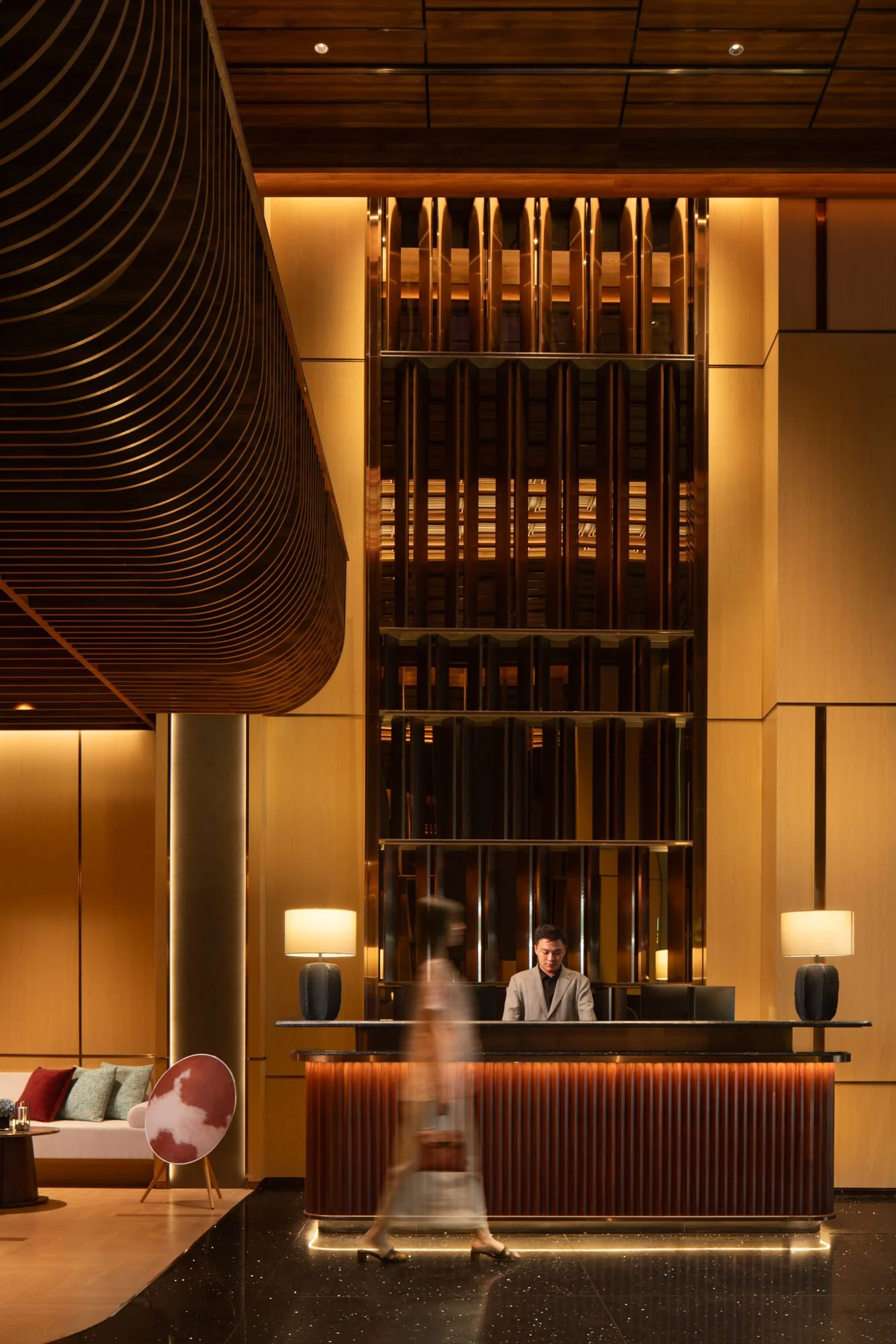  Guest walking past the reception desk in the warmly lit lobby of The Auberg Collection Hotel, featuring a towering wood feature wall and curved ceiling. 