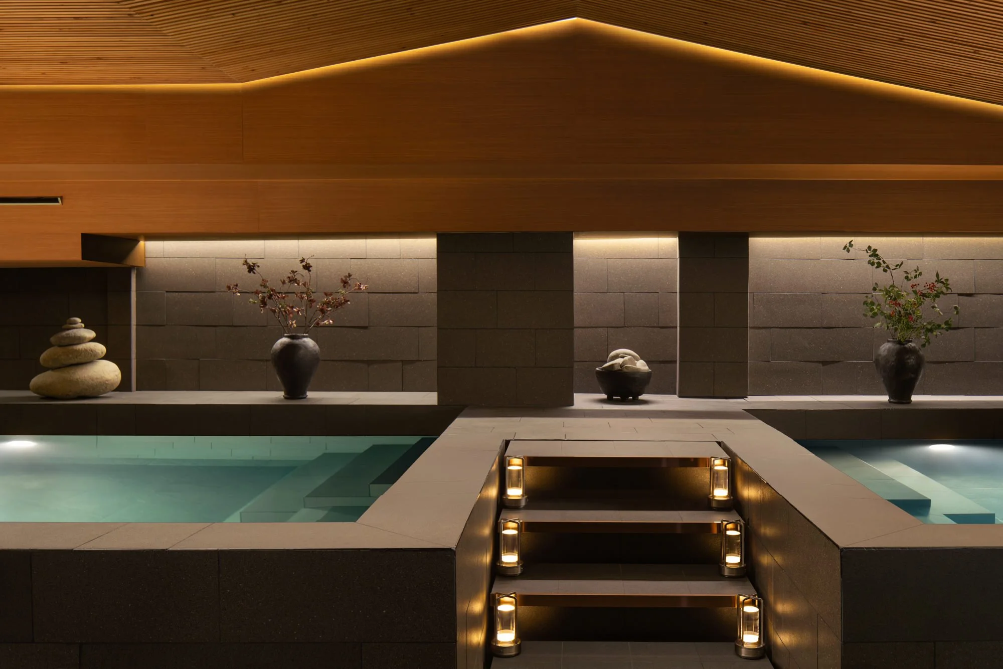  Japanese-inspired onsen at The Auberg Collection Hotel, featuring a turquoise reflecting pool, warm wood ceiling, natural stone walls, flowering branches and lantern-lit steps. 