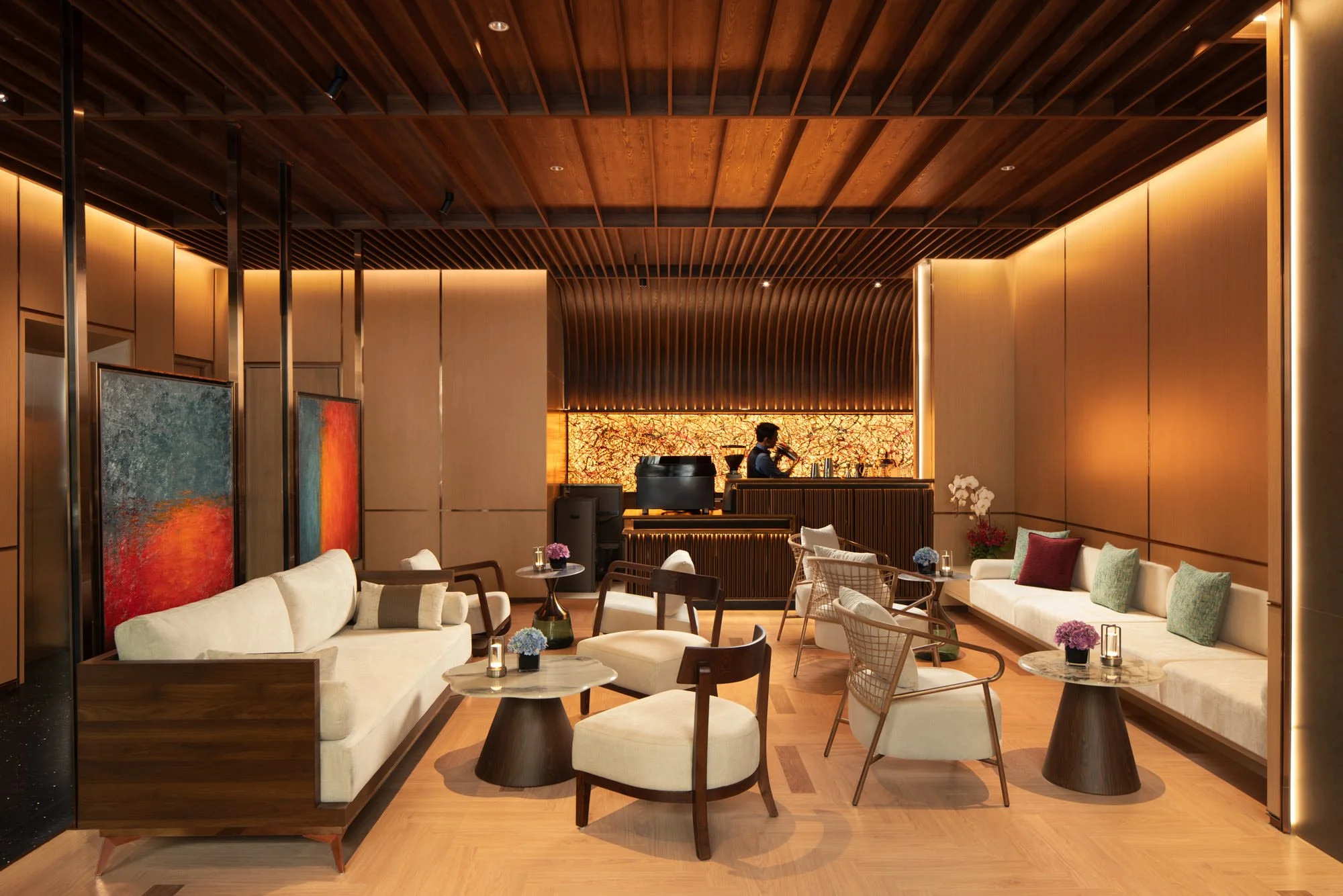  Barman shaking a cocktail behind the glowing bar at The Auberg Collection Hotel, surrounded by cream sofas, sculptural chairs and abstract artwork beneath a warm wood-slat ceiling. 