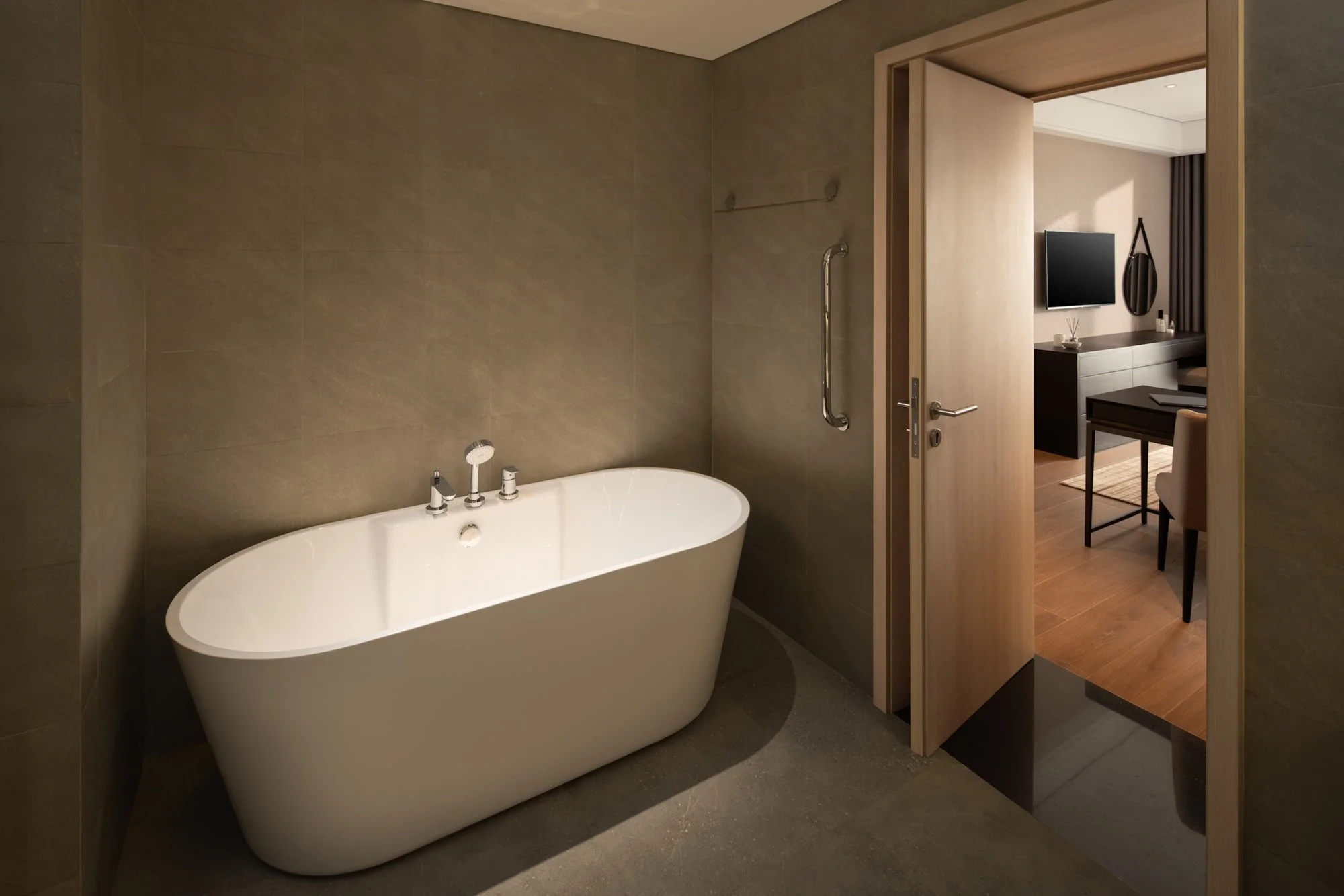  Freestanding white bathtub in the One Bedroom Executive at Modena by Fraser Vinh Yen, set within a warm, minimalist grey-tiled bathroom with an open wooden door revealing the desk and TV in the adjacent bedroom. 