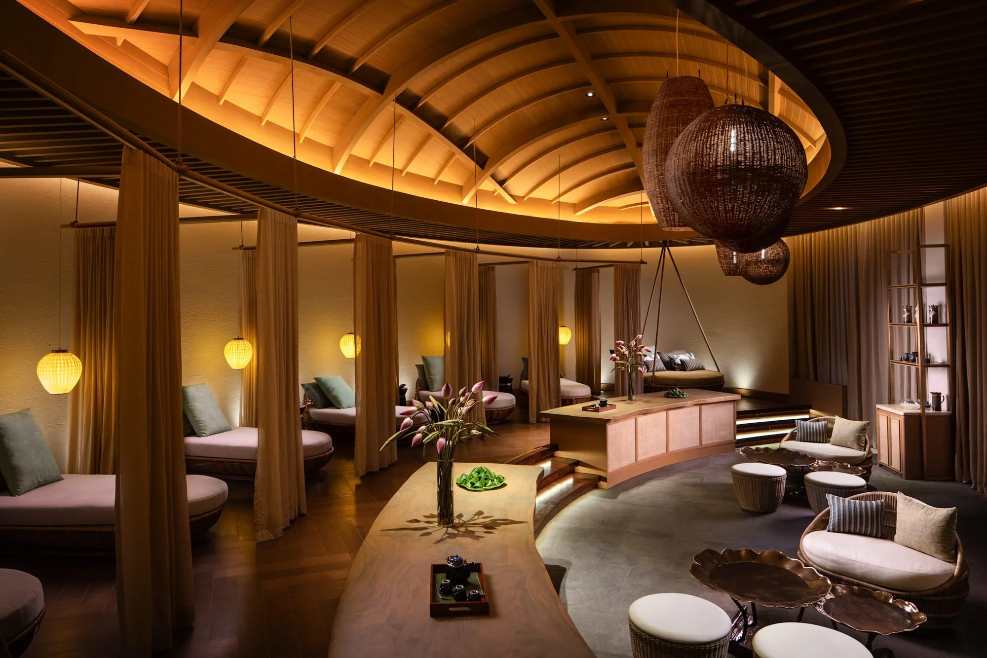  Serenity Spa foot reflexology centre at Mövenpick Resort Cam Ranh, featuring a sweeping timber ceiling, warm amber lighting, woven pendant lamps, curved seating and a decorative table adorned with flowers, tea sets and packages wrapped in banana lea