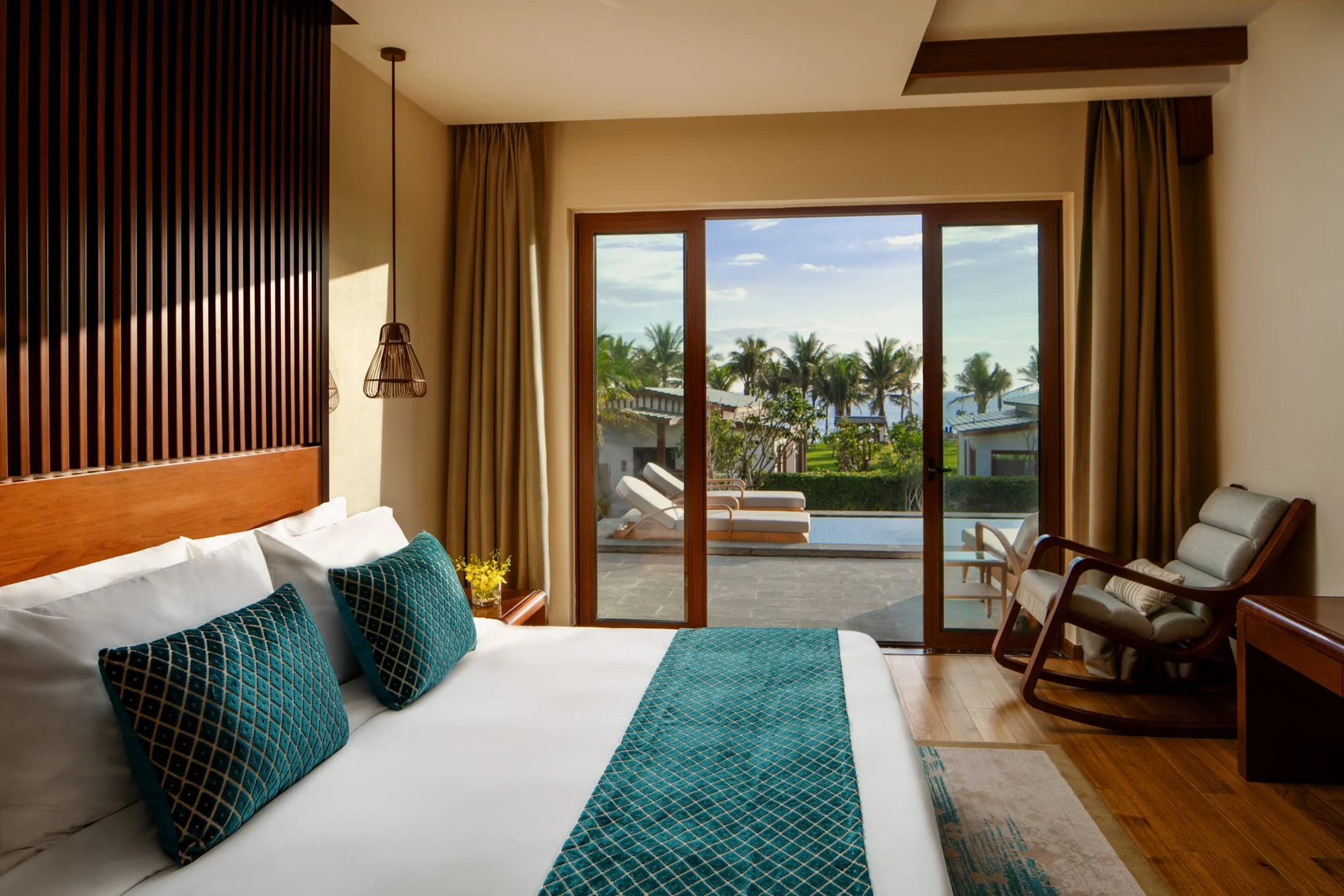  Sea View Bedroom in a modern pool villa, featuring a king-size bed with teal accents and warm wood furnishings, opening onto a balcony with lounge chairs, a private pool, tropical palms and ocean views. 