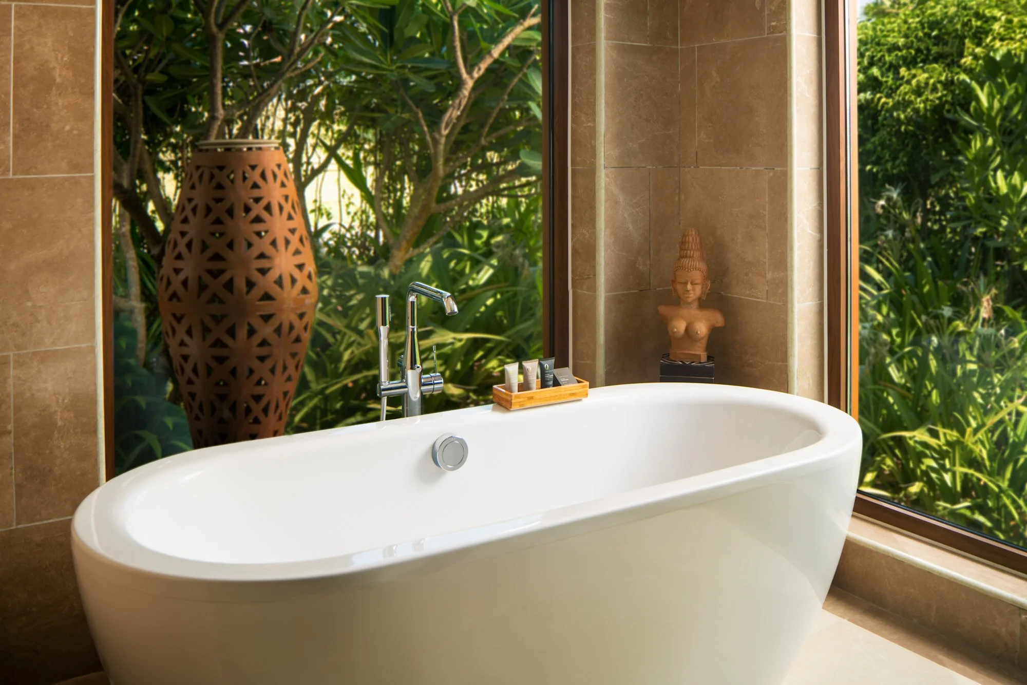  Sculptural white soaking tub beside floor-to-ceiling windows overlooking lush greenery in the Sea View One Bedroom Pool Villa at Mövenpick Resort Cam Ranh, with a carved vase and Buddha bust adding to the serene, spa-like setting. 
