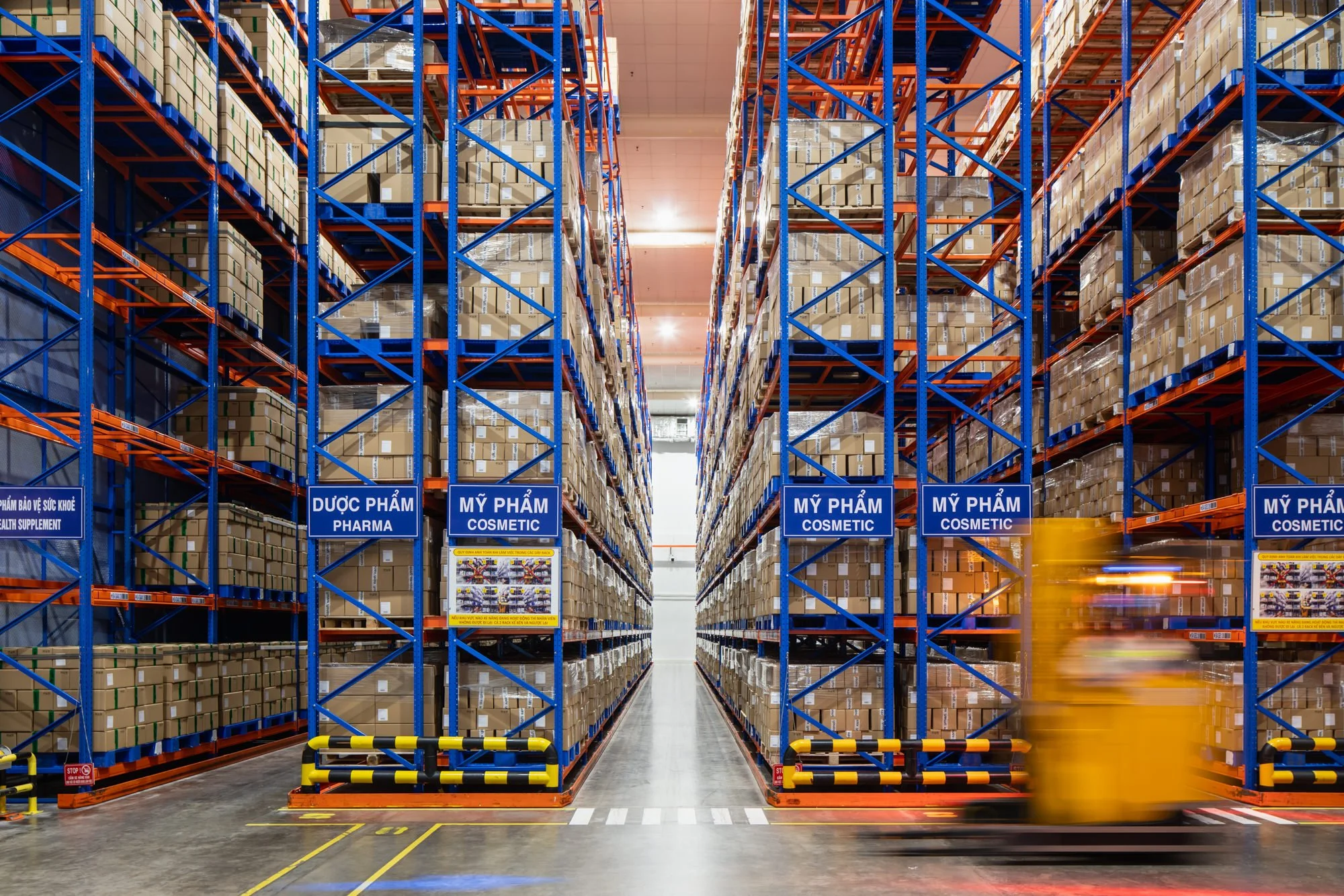  Three aisles of tall blue-and-orange warehouse racks filled with boxes of cosmetics and pharmaceuticals frame a motion-blurred forklift at the Opella manufacturing facility in Ho Chi Minh City, Vietnam. 