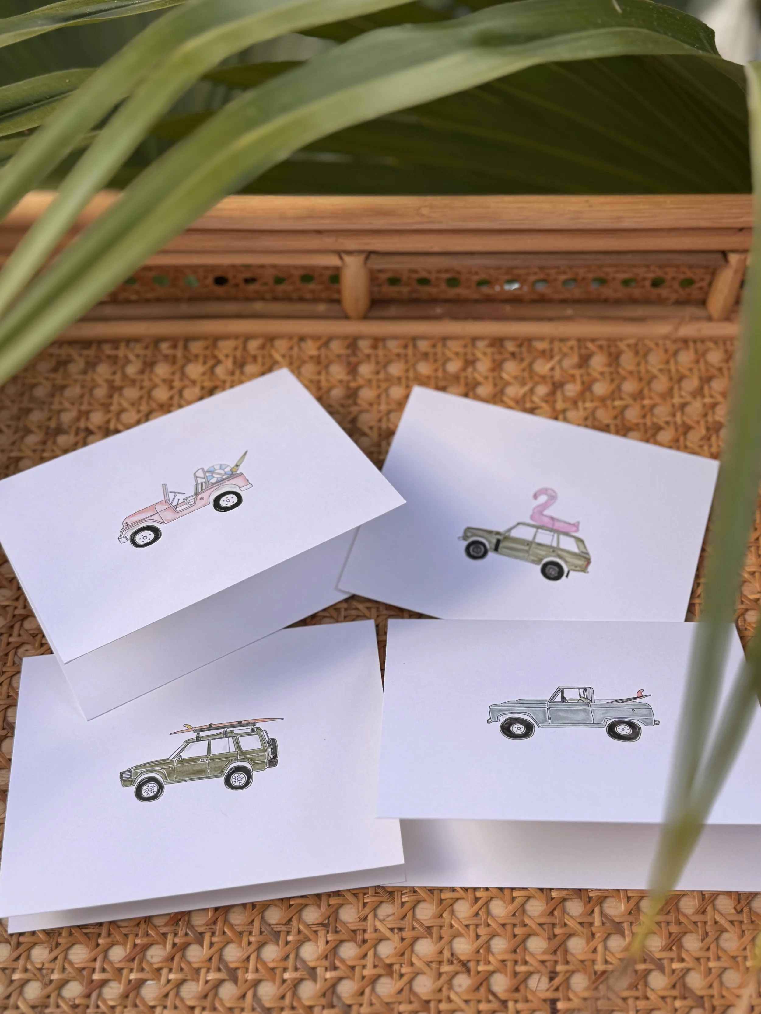 Four small paper cards with illustrations of different vehicles, including a pink jeep with a unicorn horn, a green car with a pink flamingo on top, a green off-road vehicle, and a blue pickup truck, arranged on a woven surface with large green leaves and wicker basket in the background.