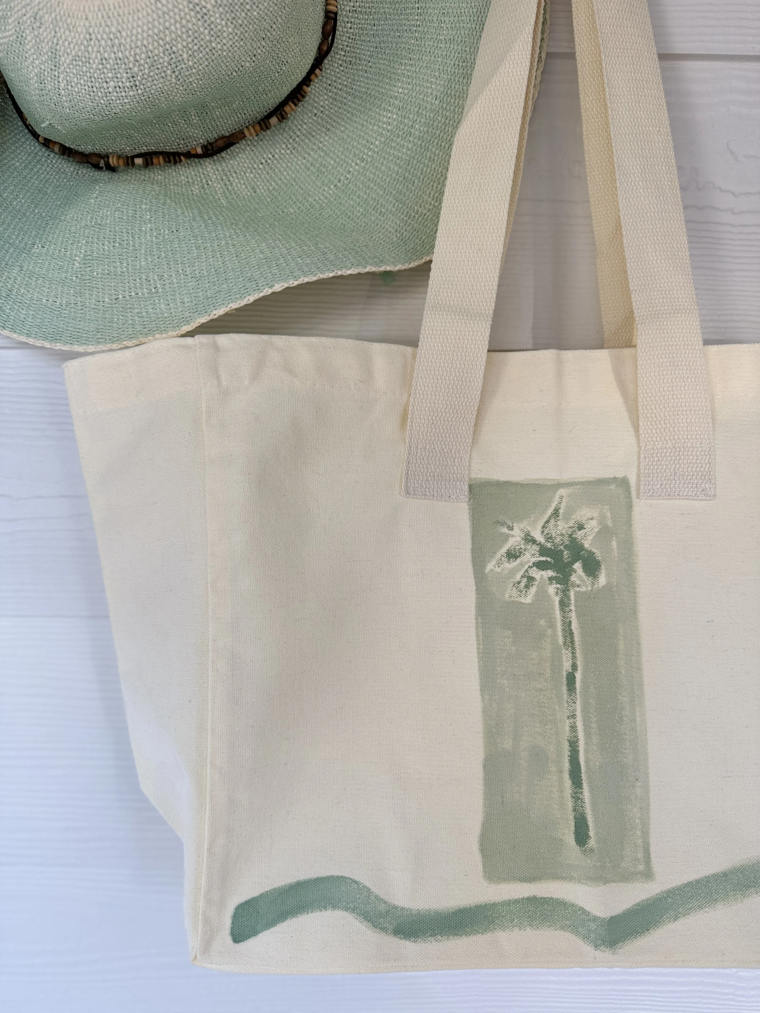 A beige tote bag with a Susie Bettenhausen palm tree graphic on it, placed on a light-colored surface, partially covered by a mint green straw sun hat with a beaded band.