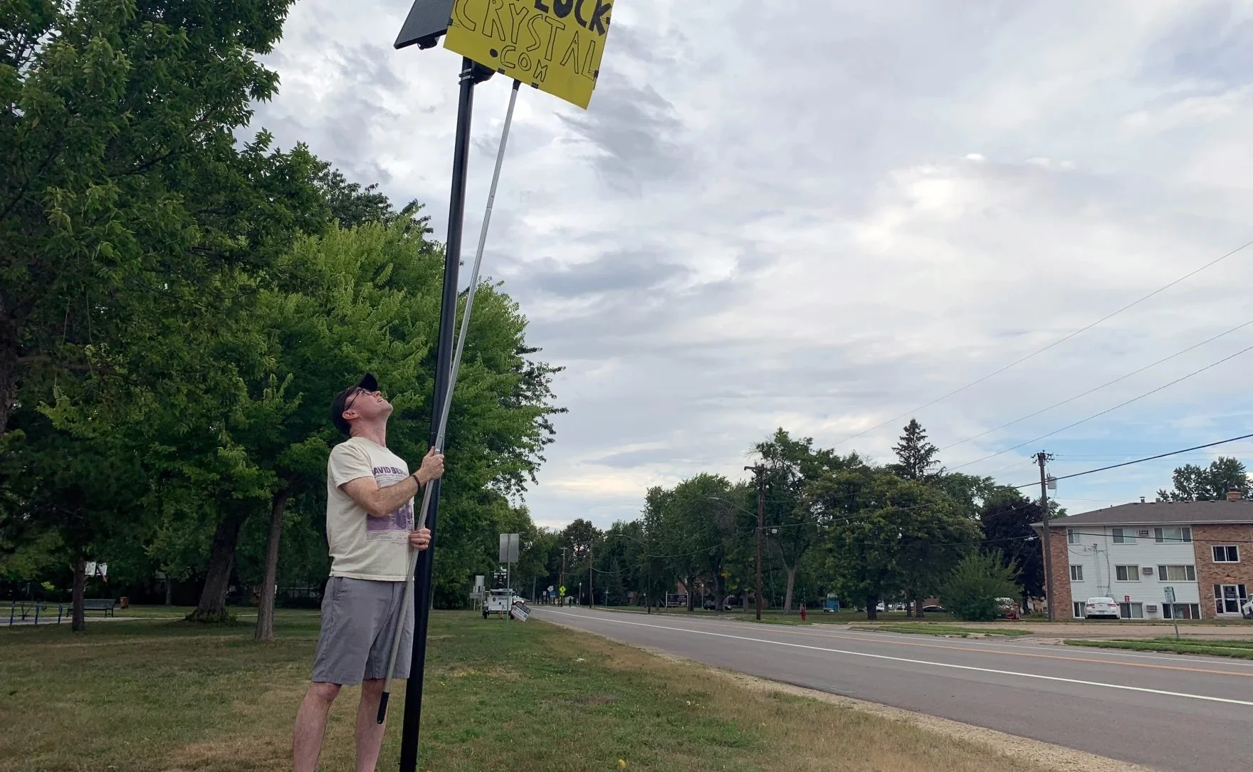 Brady O’Rourke holds a bright yellow “DEFLOCK CRYSTAL” sign in front of a roadside Flock camera in Crystal, Minnesota, blocking its view.