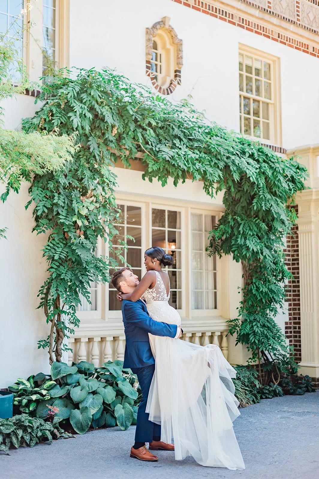 Groom lifting bride in front of the entry portico at Lairmont Manor, wisteria climbing the white stucco wall and the ornate oval window above them