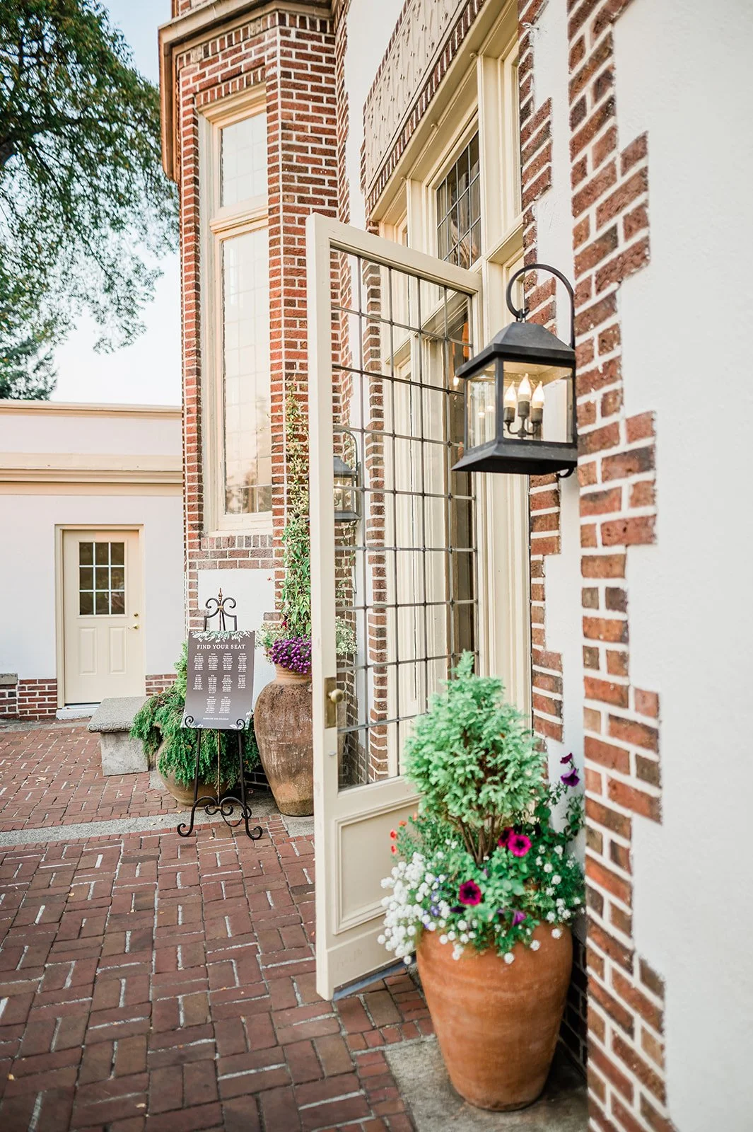 The doorway leading into Lairmont Manor from the reception side, with a seating chart on an iron stand, terracotta pots with purple flowers, and a lantern glowing against the brick exterior at dusk
