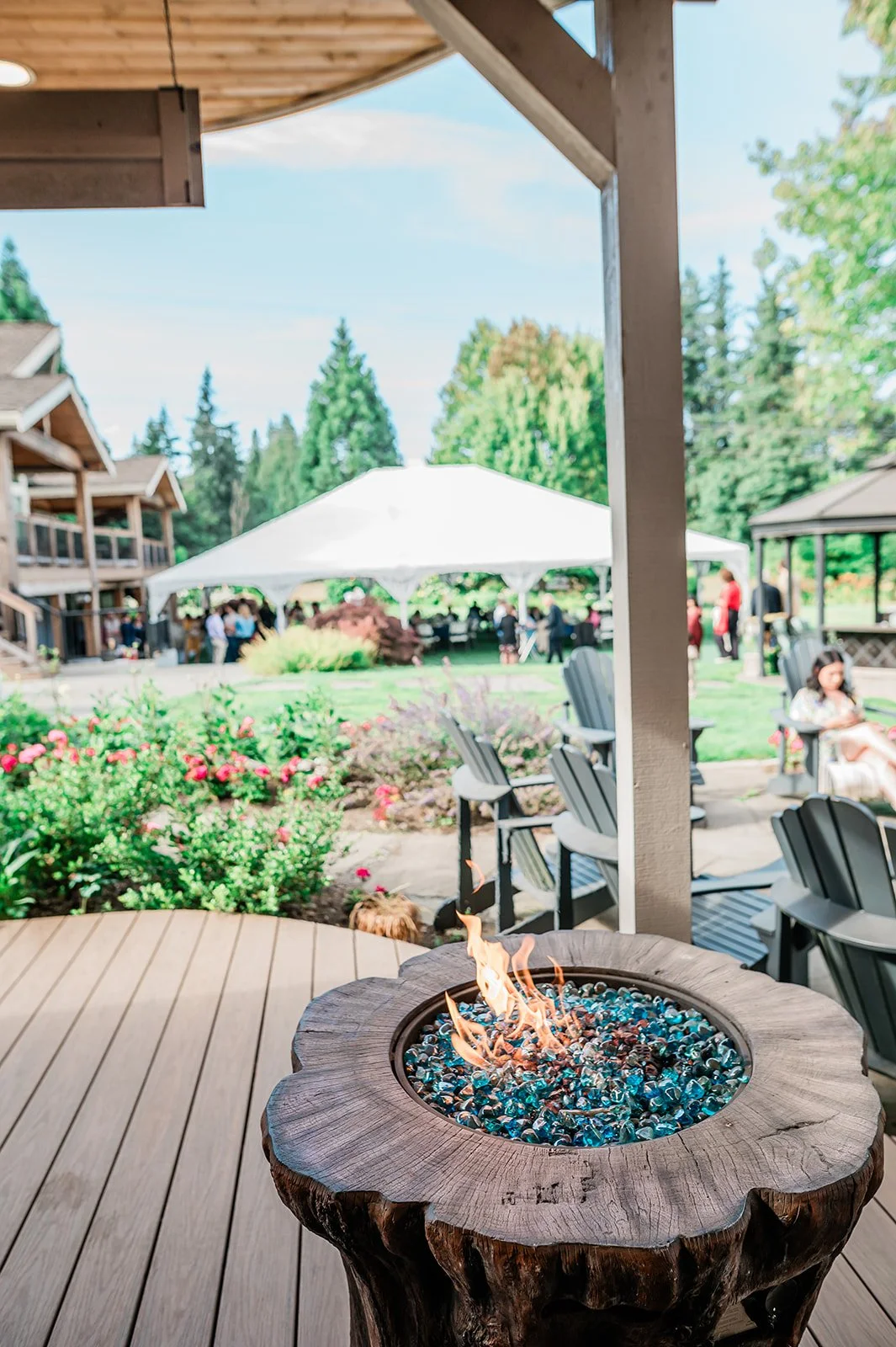 Fire pit on a wooden deck with a white reception tent and wedding guests in the background