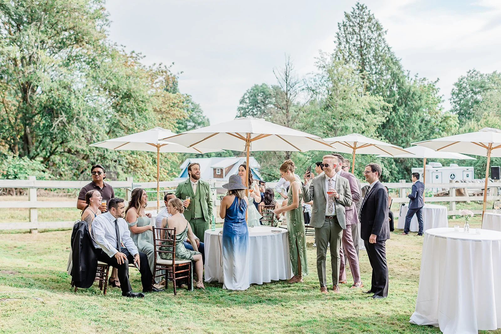 Cocktail Hour at Chateau Lill. People in formal attire socializing outdoors at a gathering with white umbrellas and round tables on a grassy area, surrounded by trees.