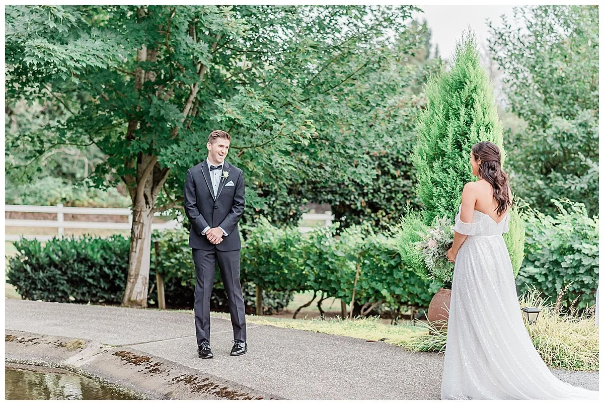 A wedding ceremony outdoors with a groom in a tuxedo and a bride in an off-shoulder white gown facing each other against a backdrop of lush green trees and bushes.