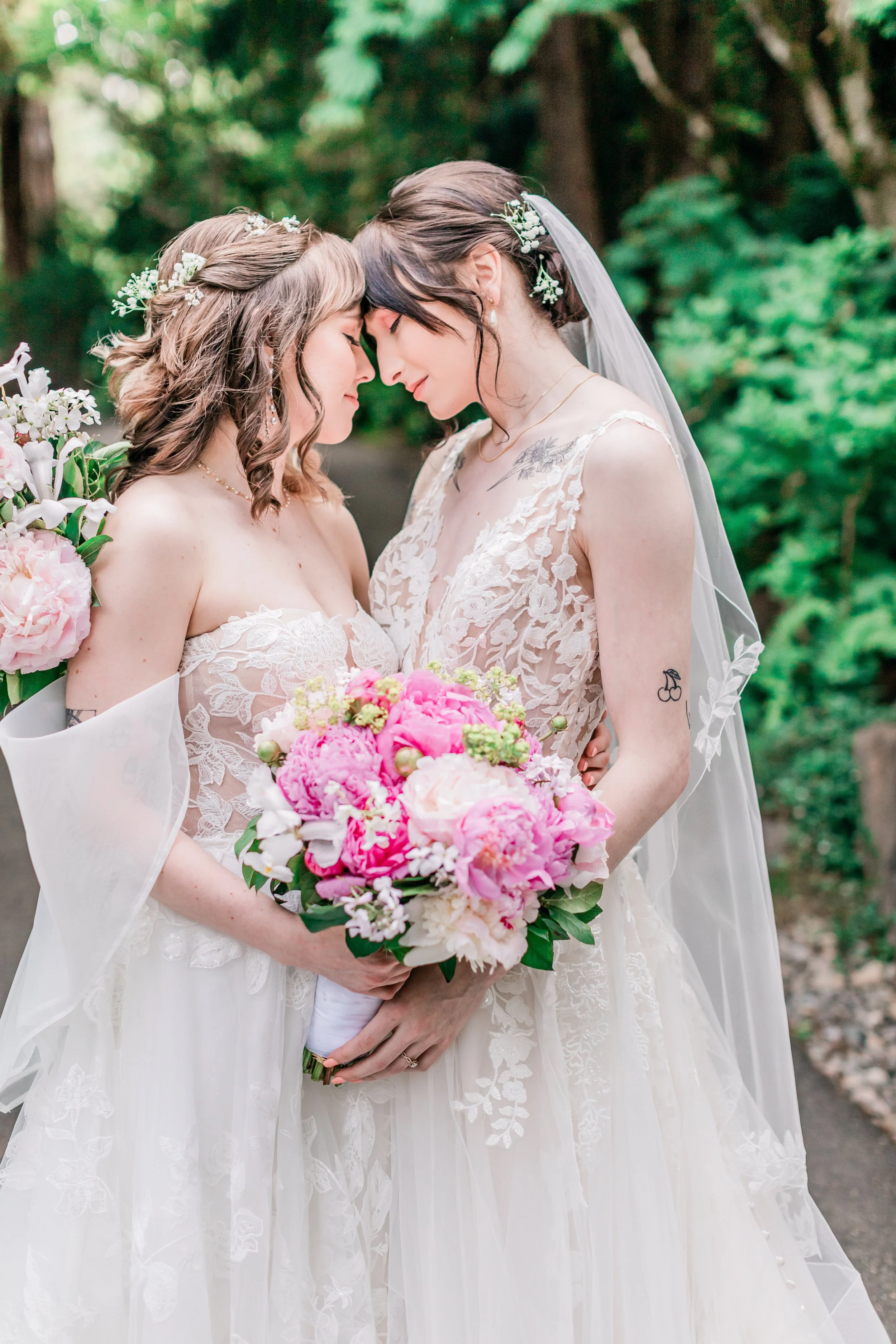 Two brides in wedding dresses holding bouquets, touching foreheads in a garden setting.