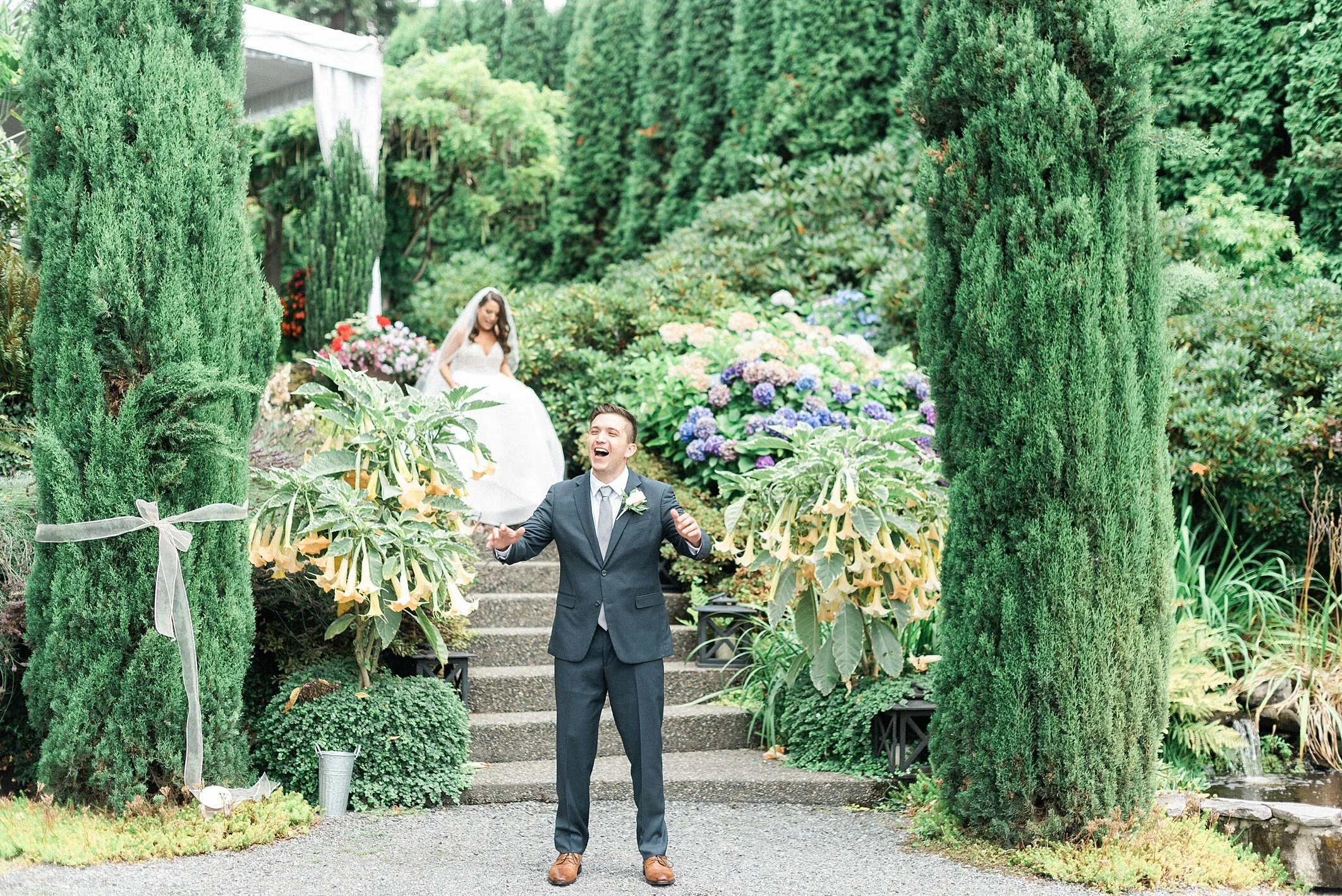 Wide shot of a wedding ceremony in the fountain garden at Lairmont Manor in Bellingham WA, with guests seated on either side of the brick aisle, the blue fountain in the foreground and the manor behind