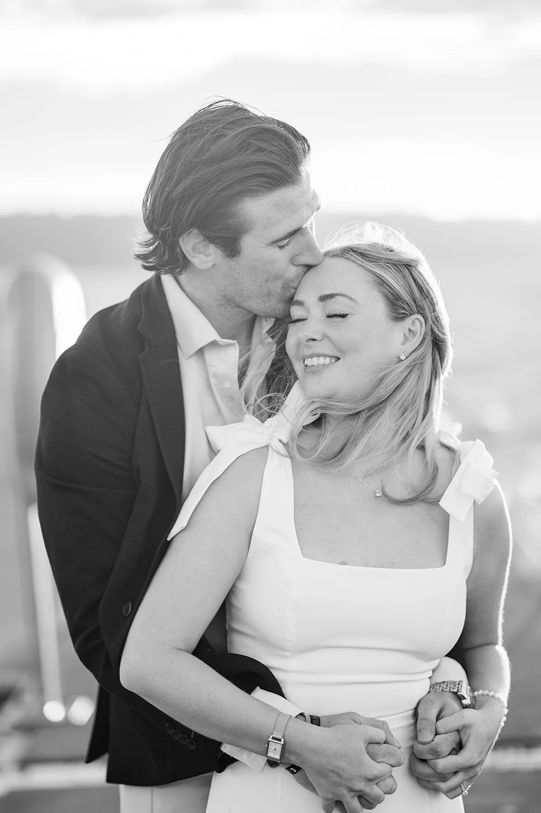 Black and white photo of fiancé kissing fiancée's cheek by the water