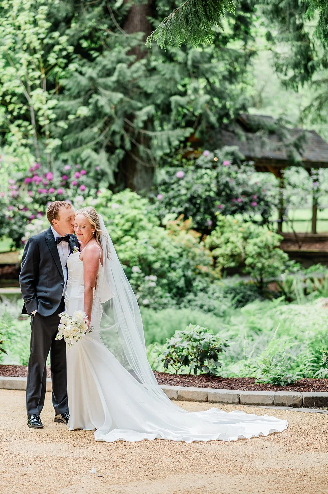 A bride and groom stand close together in a lush garden at Chateau Lill, with the groom kissing the bride on her temple. 