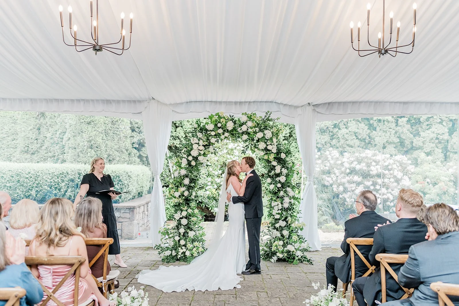 First kiss under the white floral arch during a tented ceremony at Chateau Lill