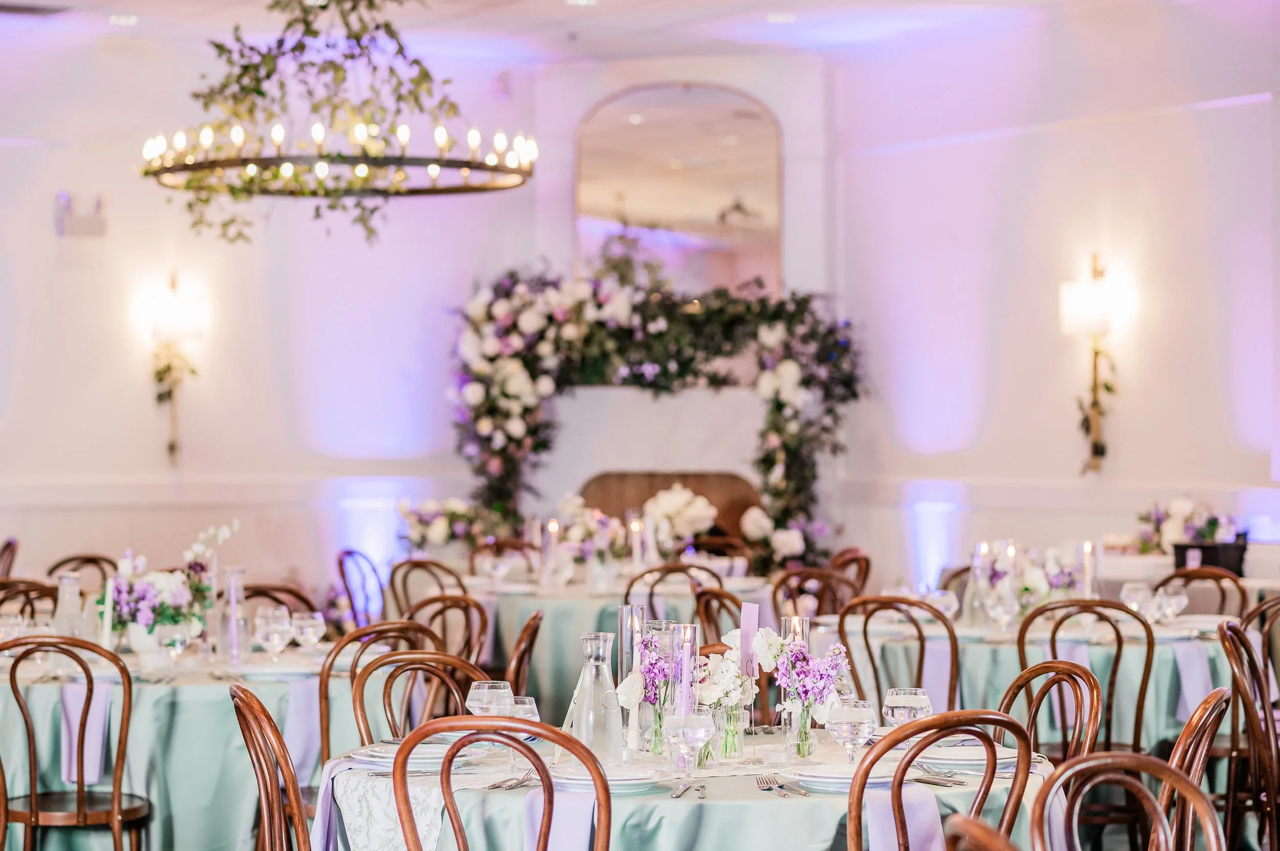 Elegant banquet hall decorated for a wedding reception with round tables covered in light blue tablecloths, floral centerpieces, and a floral arch in the background, illuminated by warm wall sconces and a chandelier.