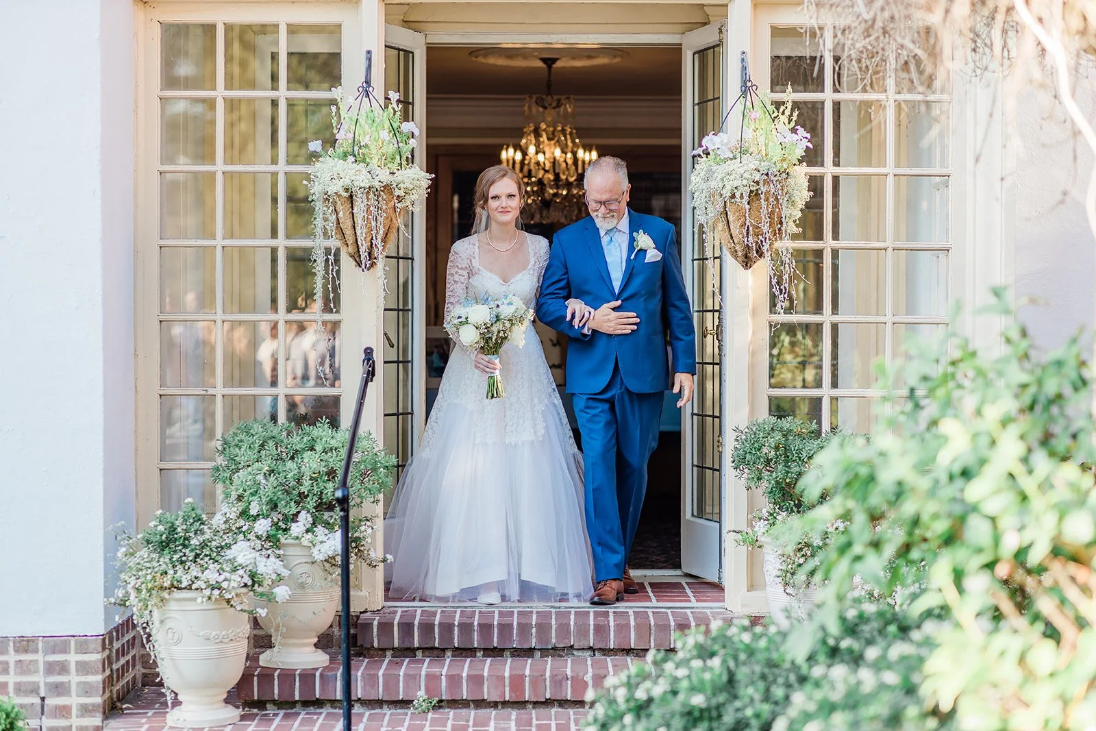Bride and her father stepping through the French doors of Lairmont Manor into the fountain garden at the start of the wedding ceremony, a chandelier glowing behind them and hanging baskets of white flowers framing the doorway