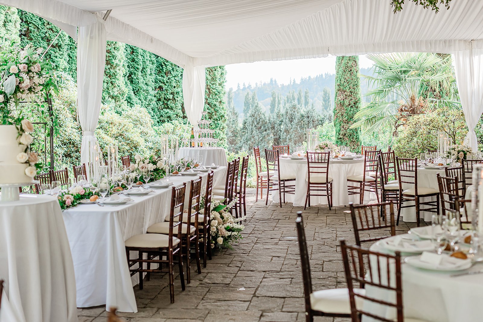 Elegant outdoor wedding reception setup at Chateau Lill with white tablecloths, floral centerpieces, and wooden chairs under a white canopy surrounded by lush greenery and trees.