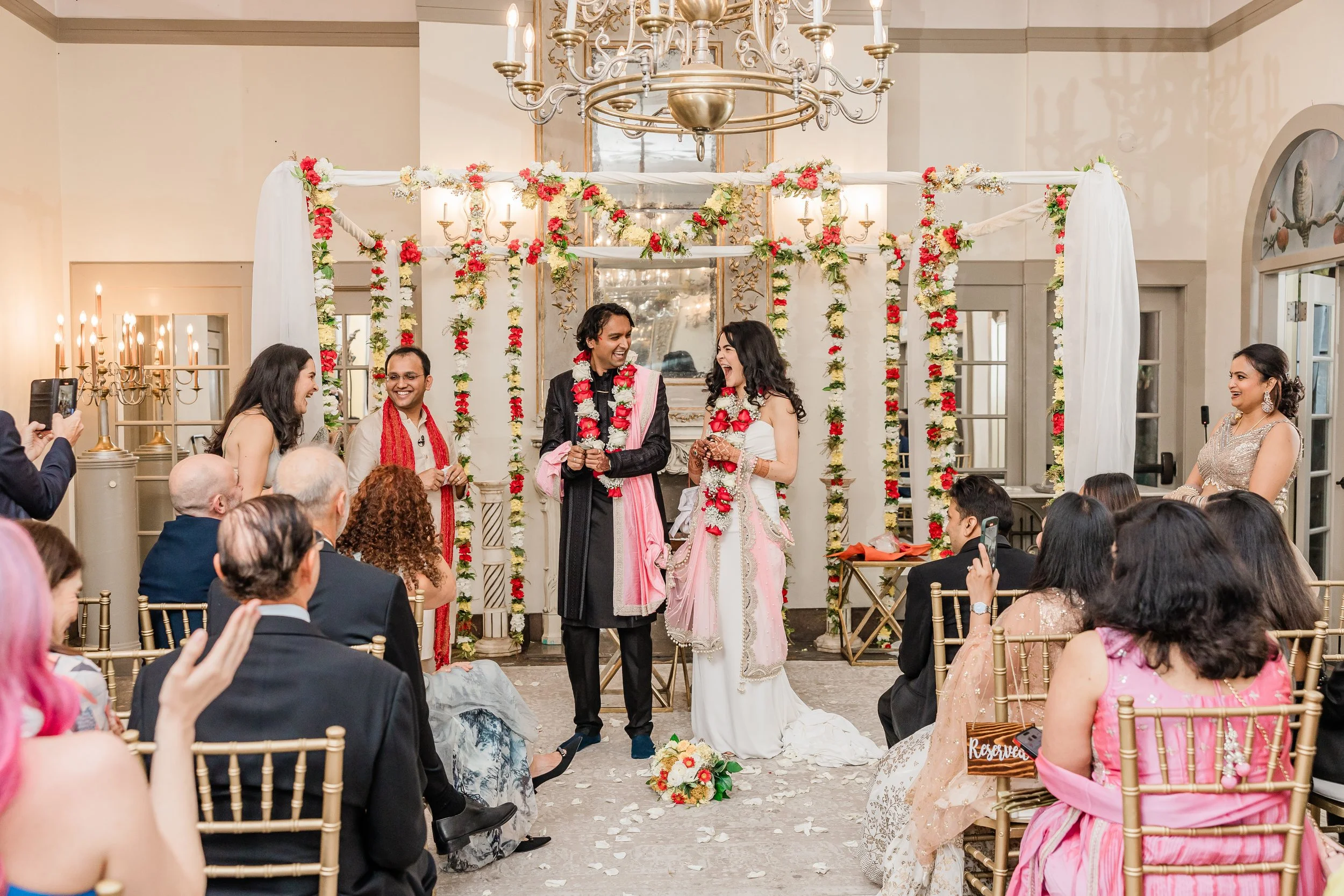 Indoor wedding ceremony under a chandelier, with guests watching the couple at the altar