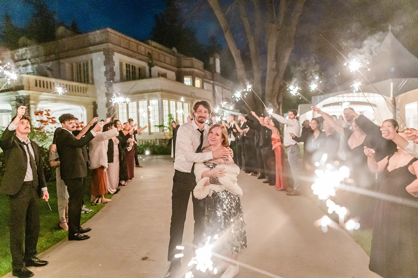 Couple embracing during a sparkler exit on the tree-lined driveway at Lairmont Manor in Bellingham at night, with the illuminated manor facade glowing in the background and guests lining both sides holding sparklers