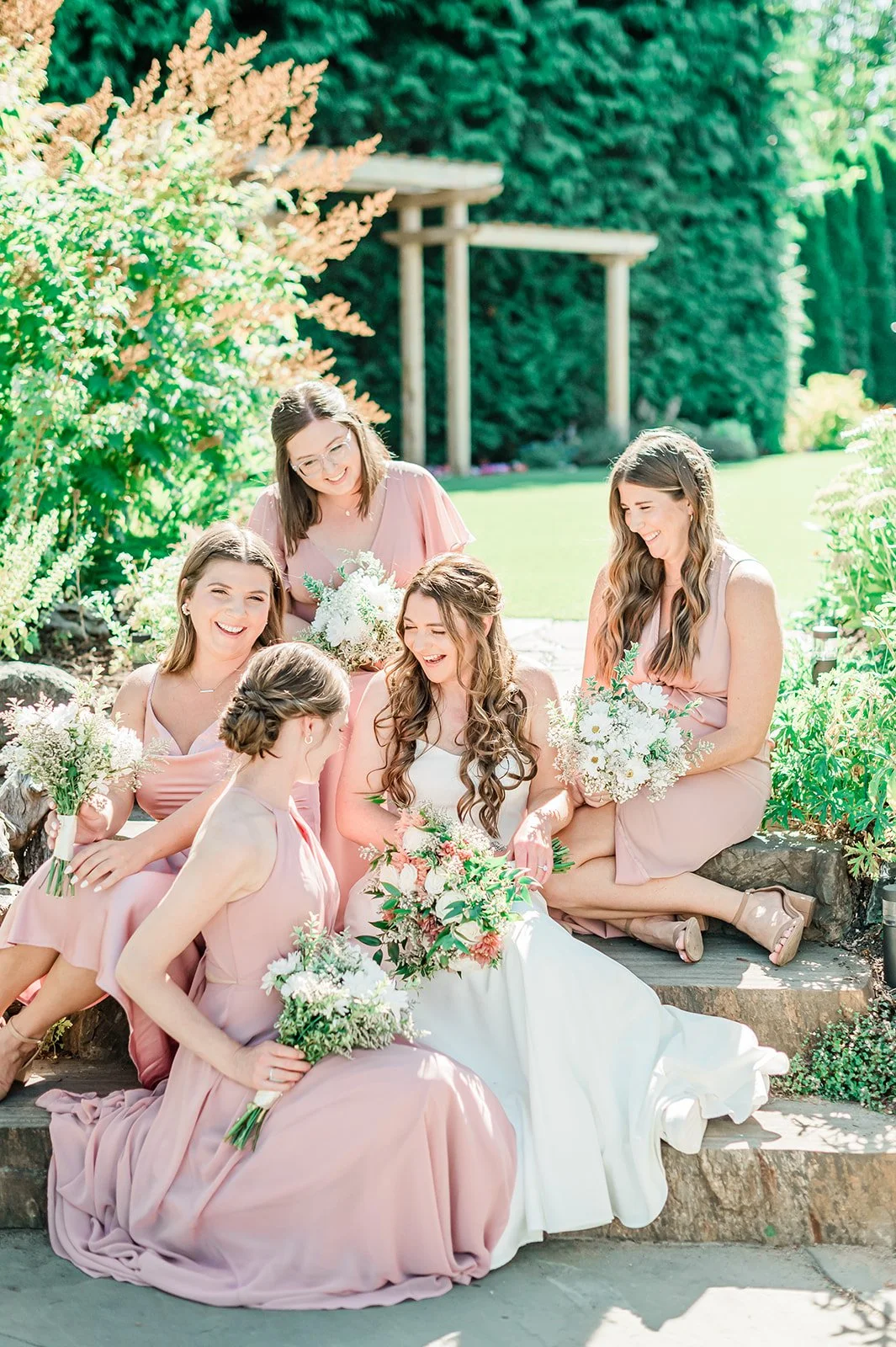Bride laughing with bridesmaids in blush pink dresses on stone garden steps under a wooden pergola