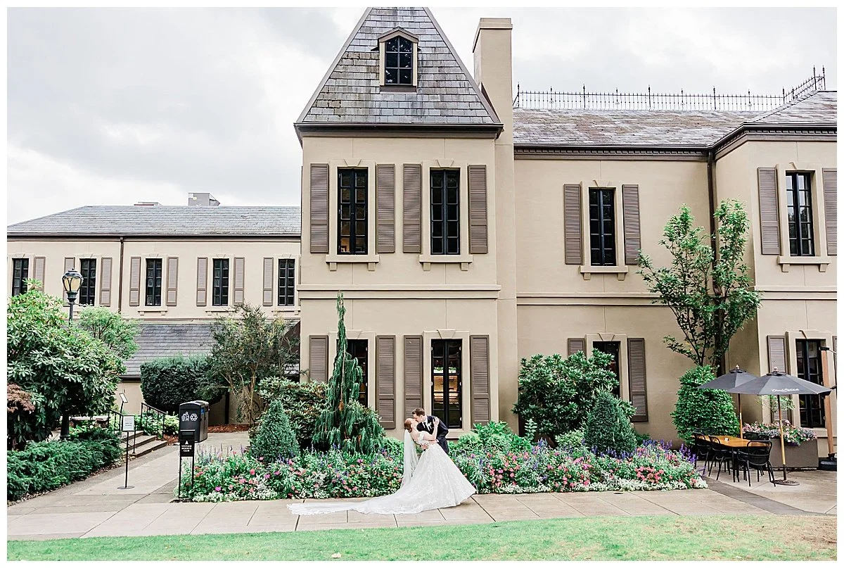 A bride and groom sharing a kiss in front of a beige mansion with brown shutters, surrounded by greenery and flowers, on a cloudy day.