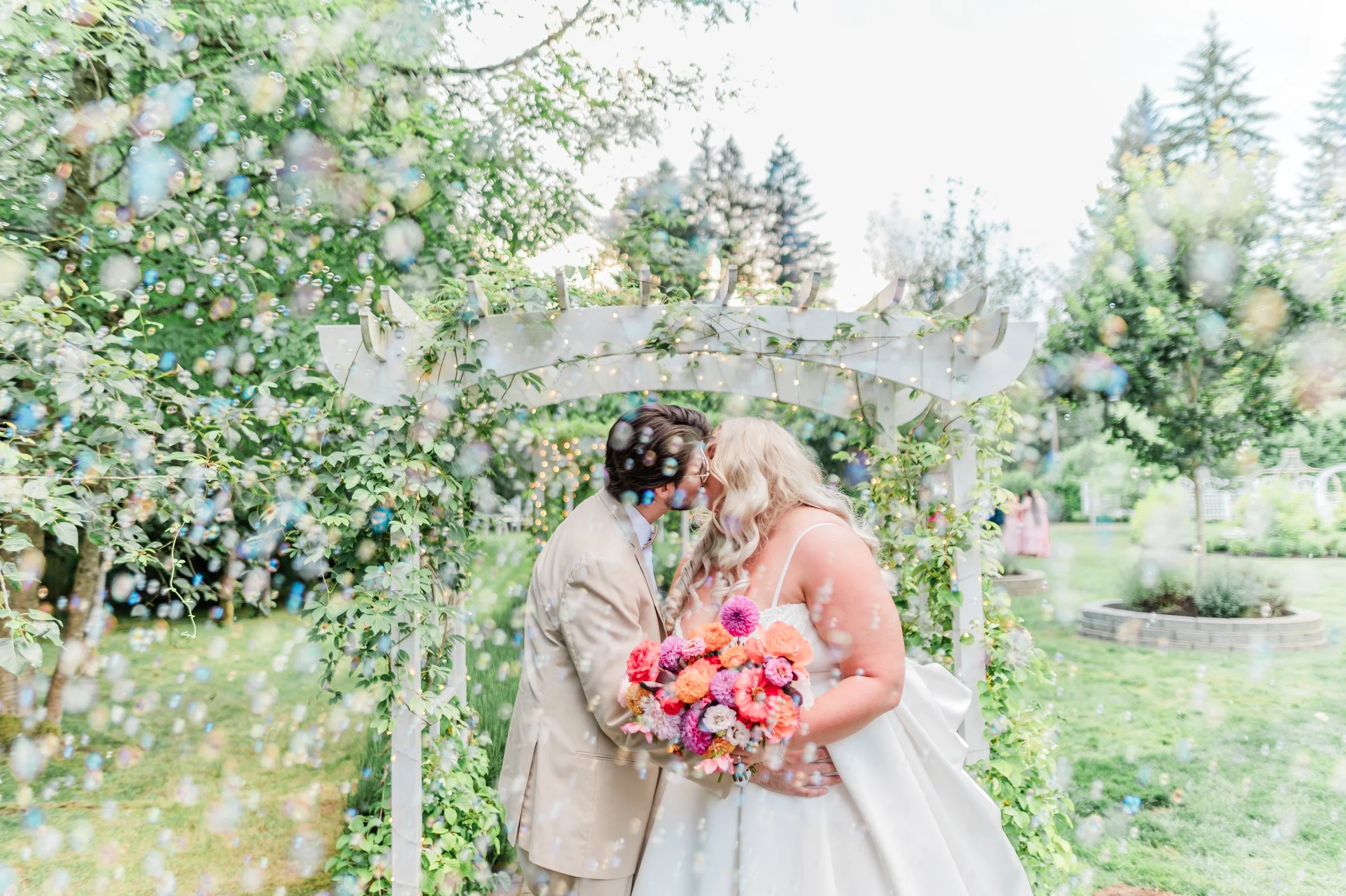 A couple sharing a kiss at a wedding ceremony outdoors, with greenery and trees in the background, decorated with string lights and an arch, while colorful flowers are held by the bride.