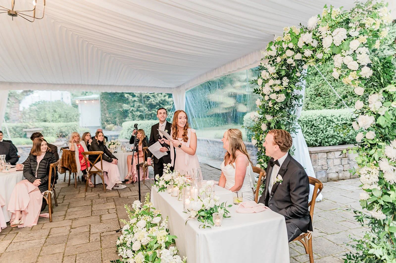 Maid of honor giving a toast beside the sweetheart table under the floral arch