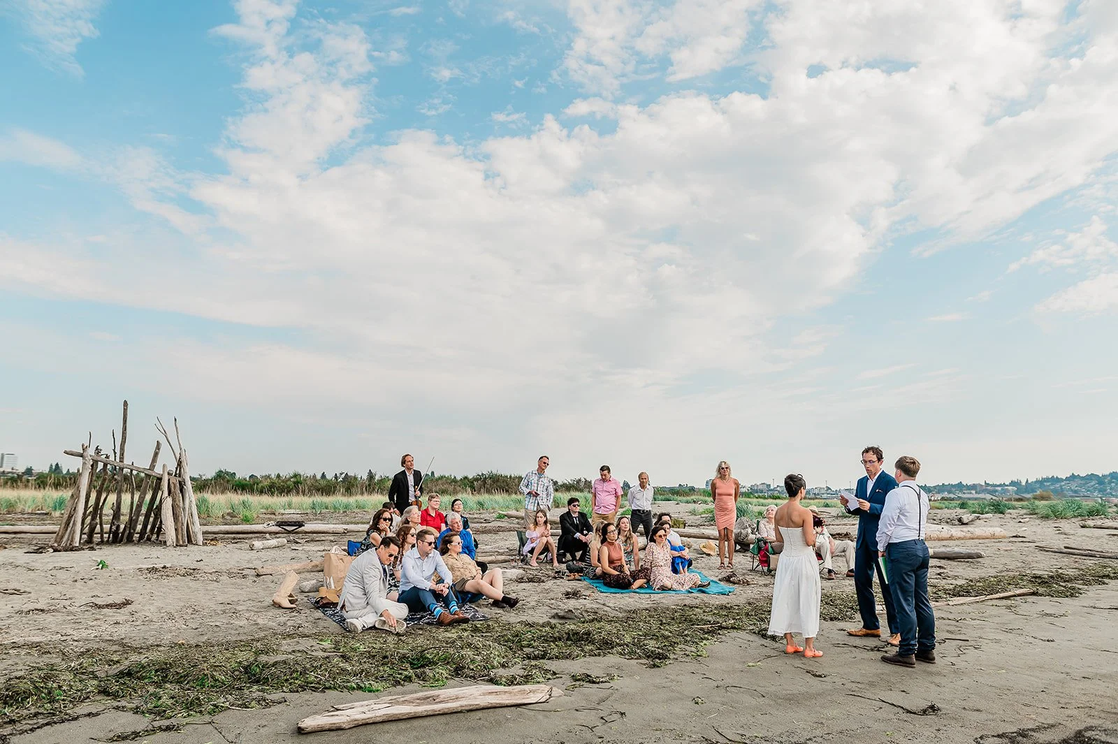 Intimate outdoor wedding ceremony on Jetty Island Everett Washington with guests seated on blankets and driftwood under a Pacific Northwest sky