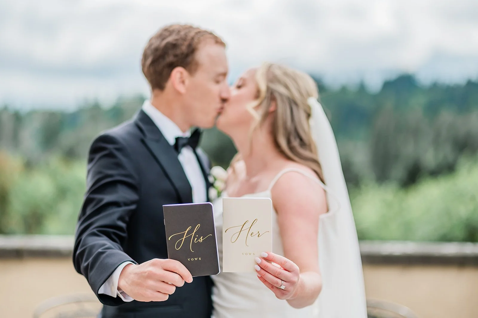 Couple kissing while holding His and Hers vow books on the Chateau Lill balcony