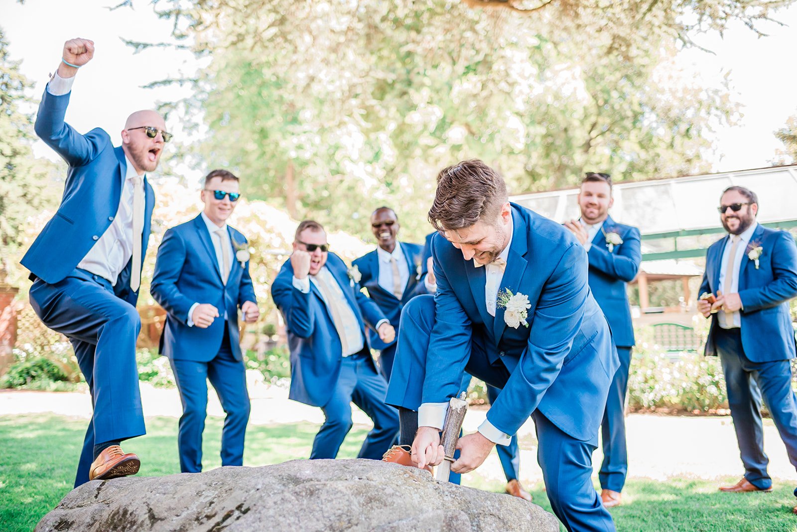 Groom pulling the sword from the stone at Lairmont Manor while his groomsmen celebrate wildly behind him, the estate's giant evergreen tree visible in the background