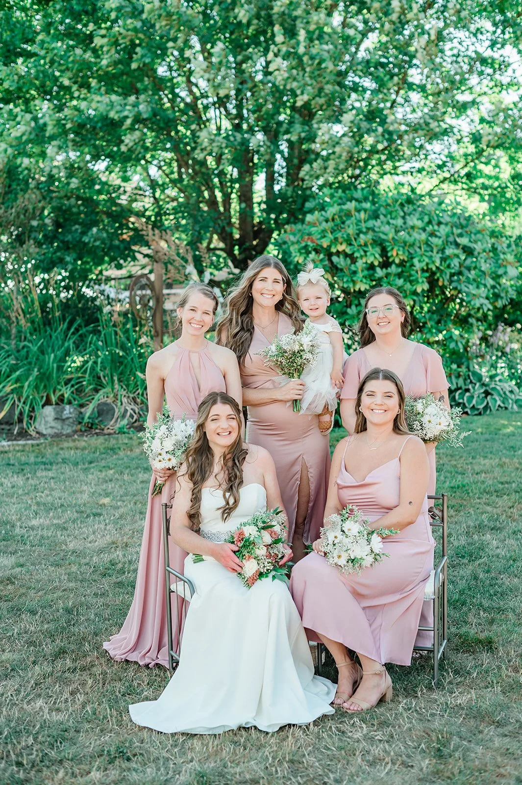 Bride seated with bridesmaids in blush pink dresses, one holding the flower girl, in the garden at The Carey Gardens