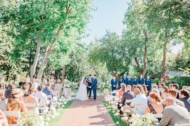Outdoor wedding ceremony with bride and groom exchanging vows under trees, guests seated on either side, sunny day with blue sky.