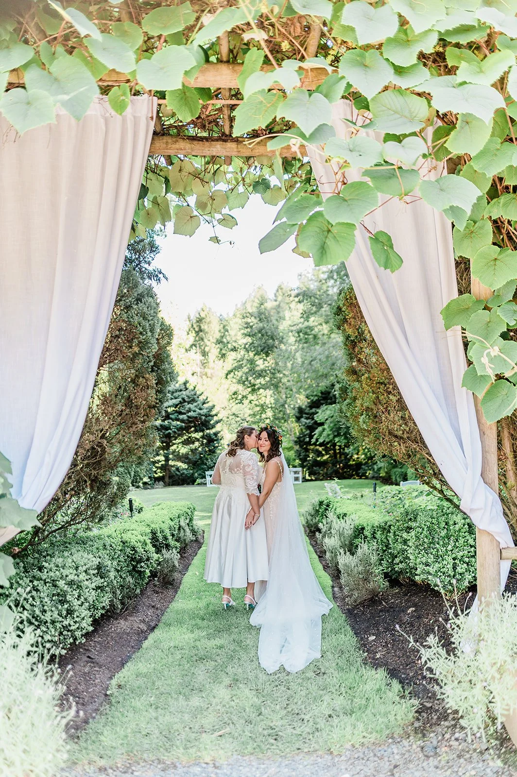 Two brides framed by vines and a hedge path at Jardin del Sol in Snohomish
