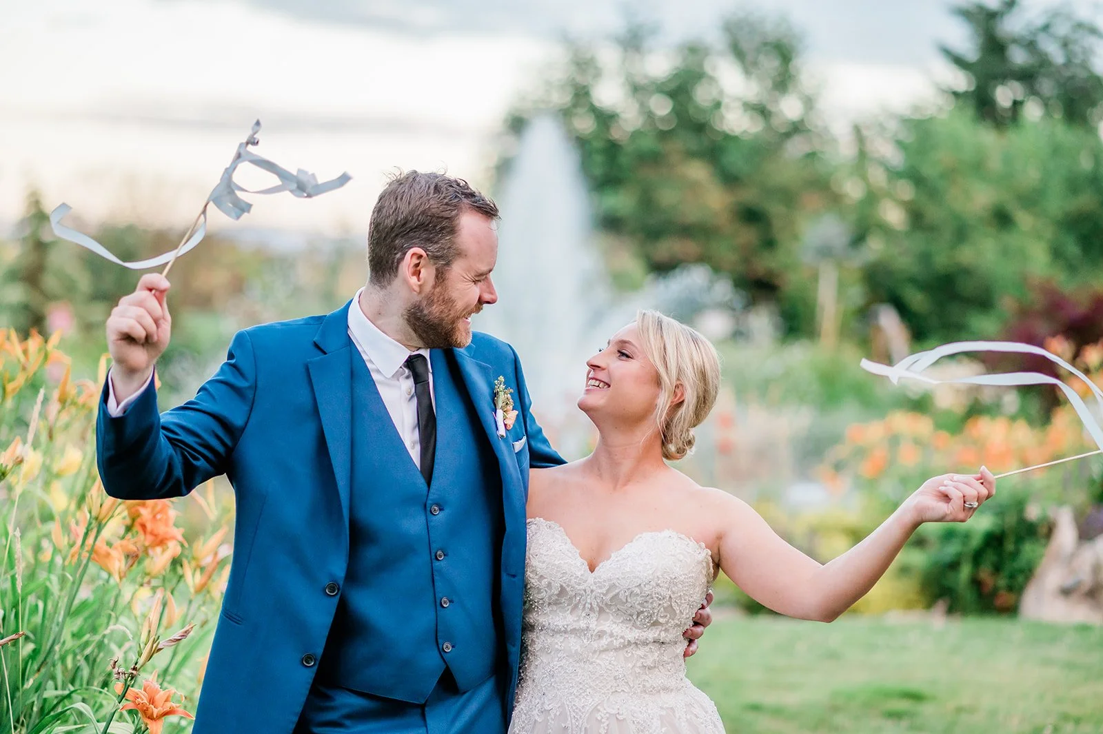 A bride in a white wedding dress holding a bouquet, and a groom in a black tuxedo, in a lush garden setting with plants and trees, during a wedding or engagement photoshoot.