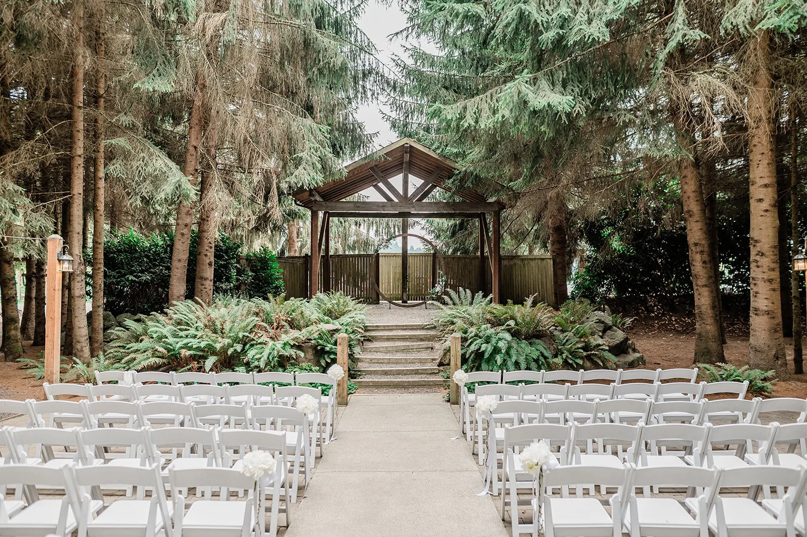 Forest wedding ceremony site with a wooden arbor and white chairs beneath towering pines at Woodland Meadow Farms
