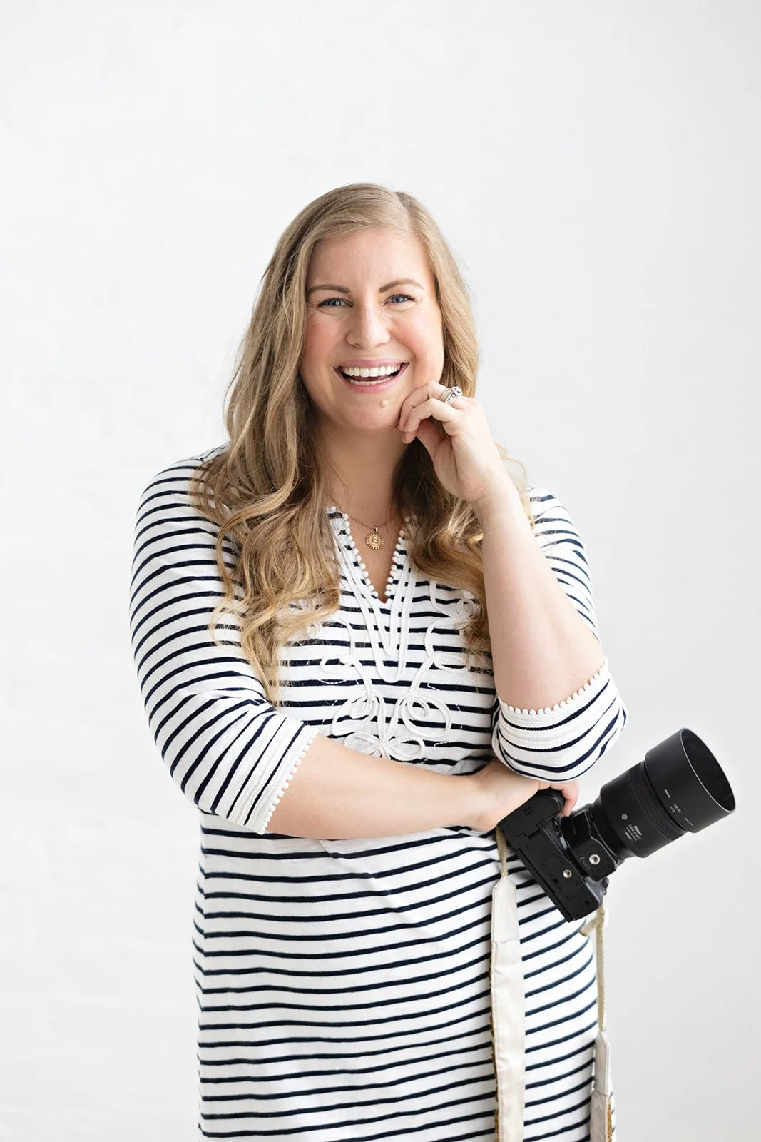 A woman with long wavy blonde hair, smiling, wearing a black and white striped dress with a camera hanging from her neck, standing against a plain white background.