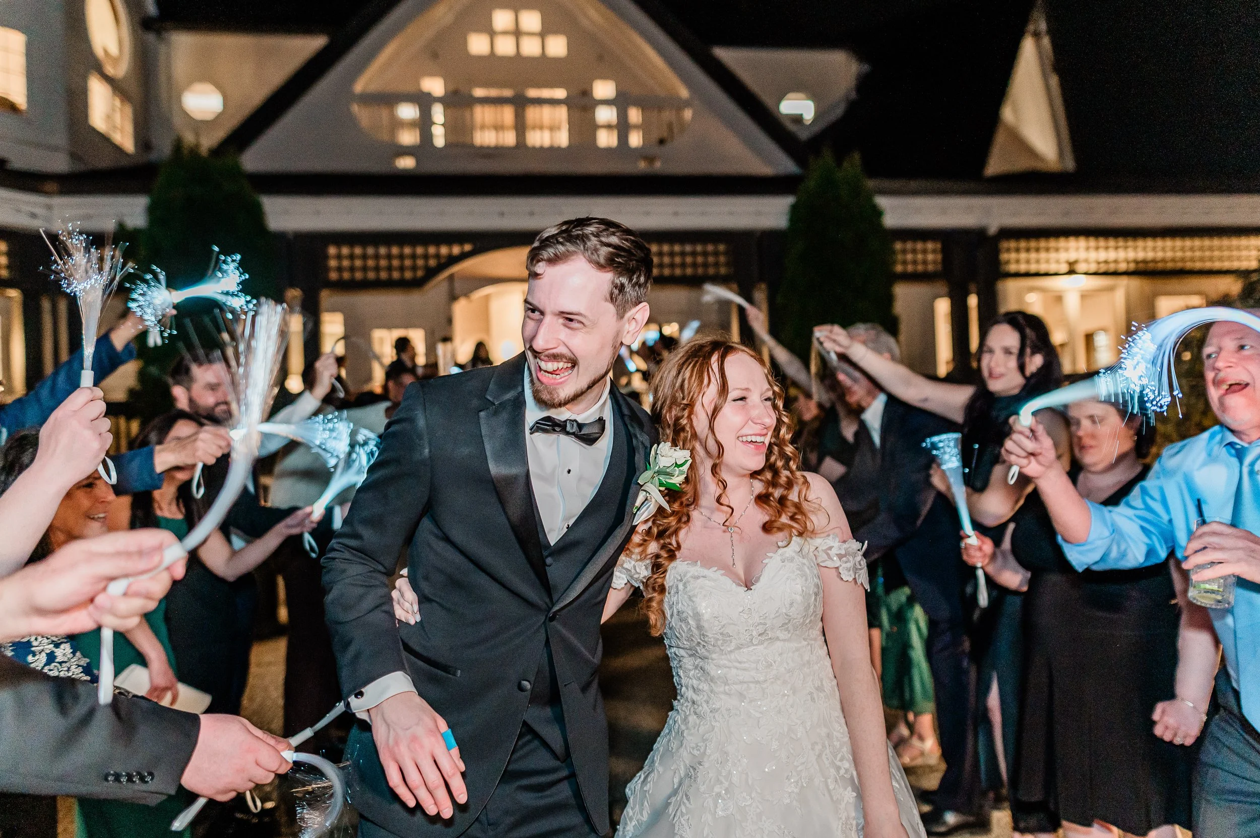 A newlywed couple, the groom in a black tuxedo and the bride in a white lace wedding dress, joyfully walking through sparklers held by friends and family at night outside a large house.